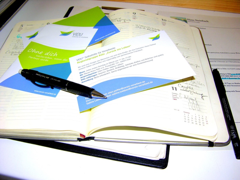 Geöffnetes Terminbuch auf welchem drei Flyer und zwei Stifte liegen