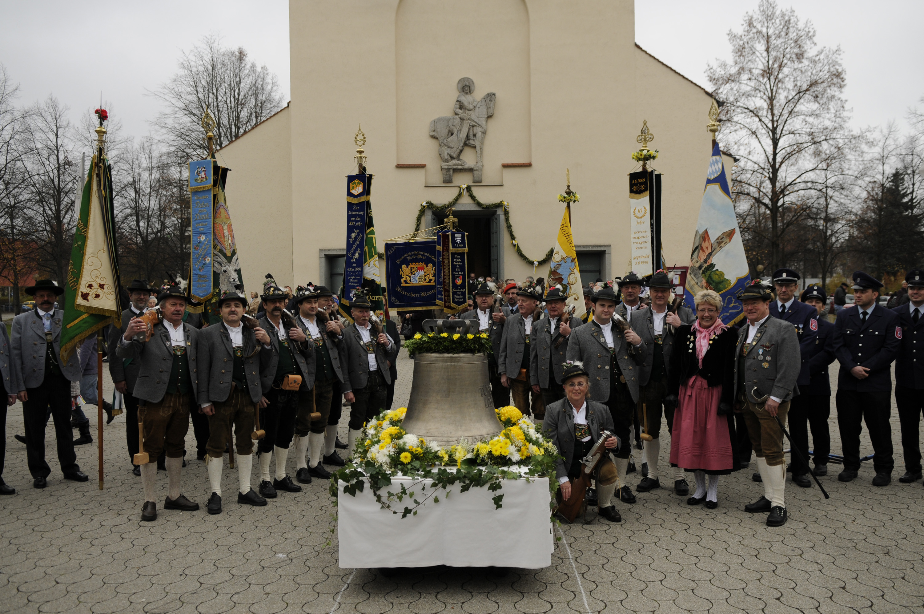 Kirchenglocke auf einem Wagen umringt von Männern in Tracht und Fahnen. Im Hintergrund das Kirchenportal.