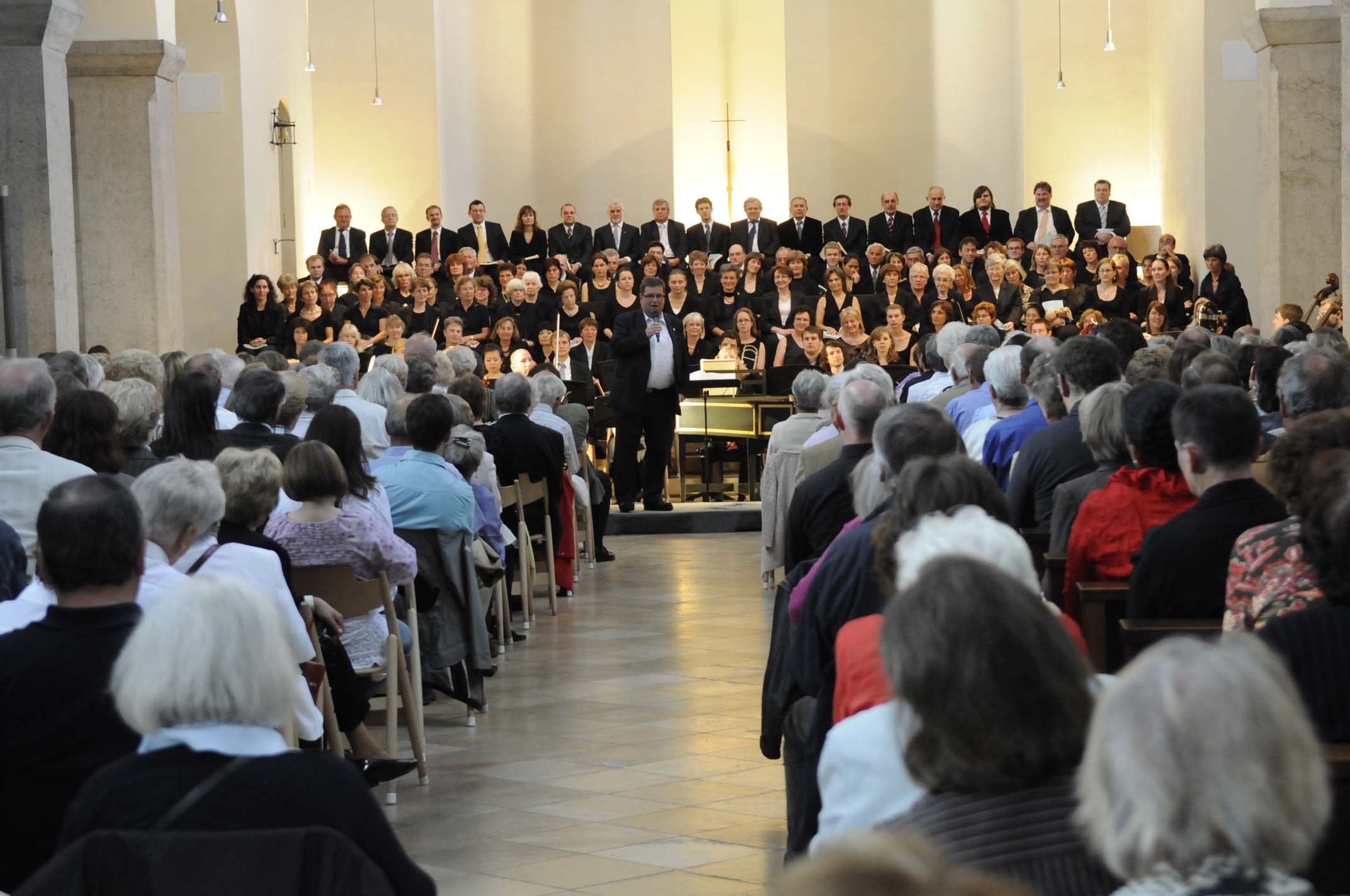 Kirche innen mit vollbesetzten Bänken und einem großen Chor mit Orchester im Altarraum. Pfarrer Cambensy macht eine Ansage