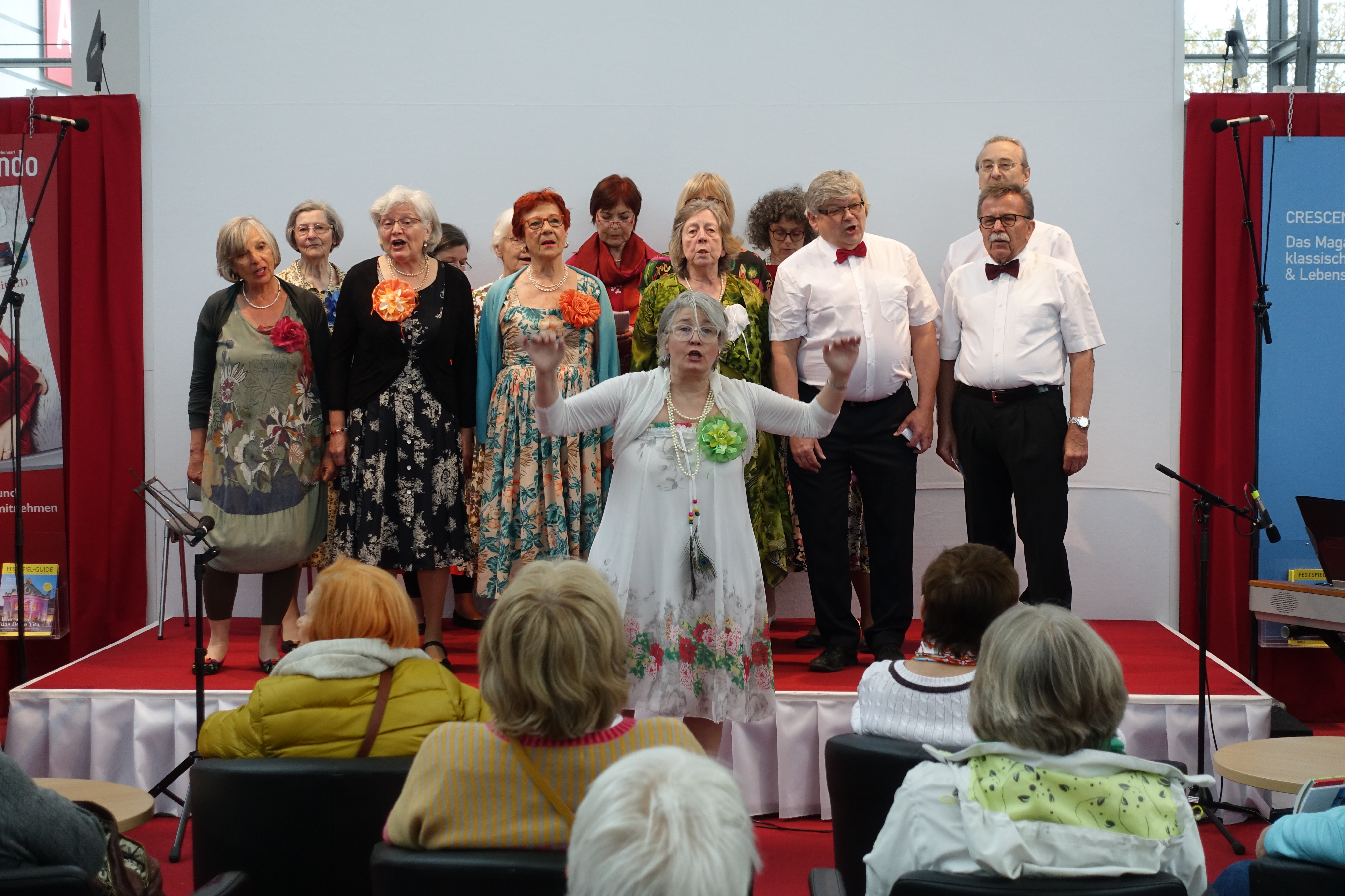 Sängerinnen und Sänger auf der Bühne singend. Rose animiert das Publikum.