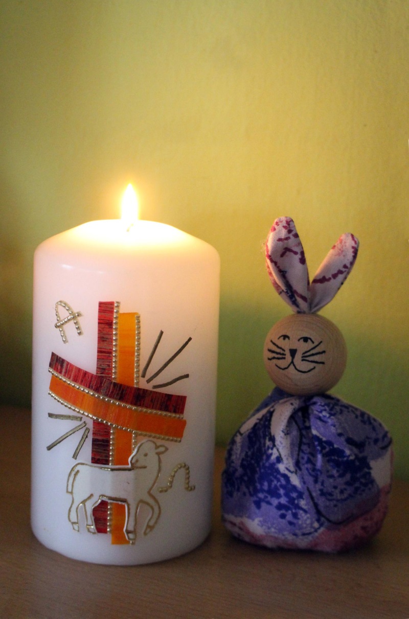 Brennende Osterkerze mit einem Kreuz aus rot und orangen Bändern verziert, daneben ein Osterhase im lila Gewand