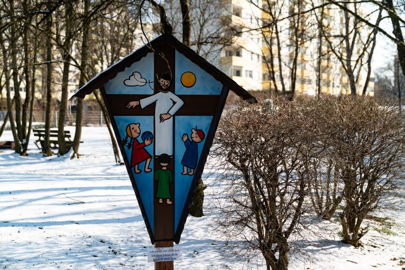 "Wegkreuz" im verschneiten Garten, darauf gemalt: Jesus spielt mit den Kindern