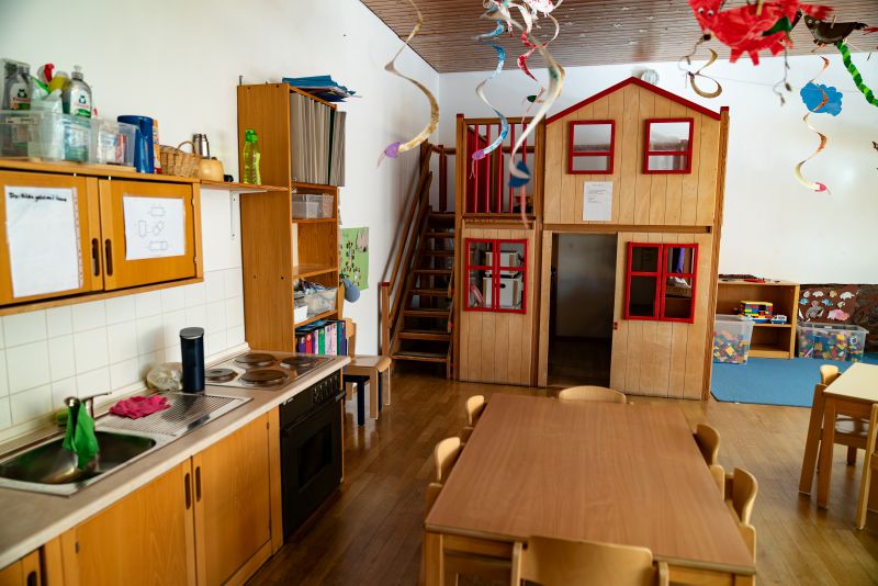 Küchenzeile und hölzernes Spielhaus mit roten Rahmenfenstern im Gruppenraum der Kita St. Hildegard.