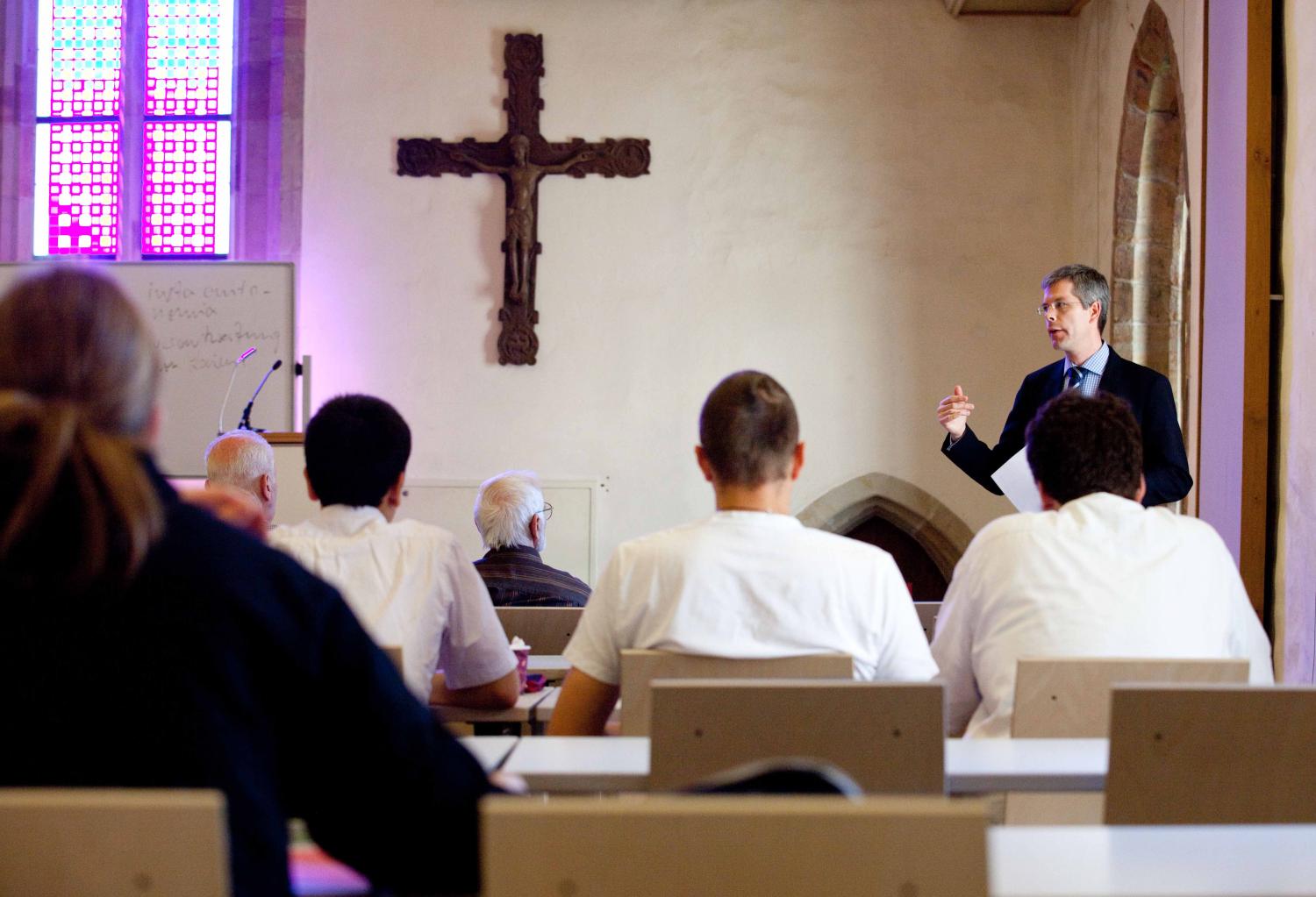 Mehrere Köpfe mit kurzen Haaren von jüngeren und älteren Menschen, sitzend, gegenüber stehend ein Mann, der vorträgt, großes Wandkreuz