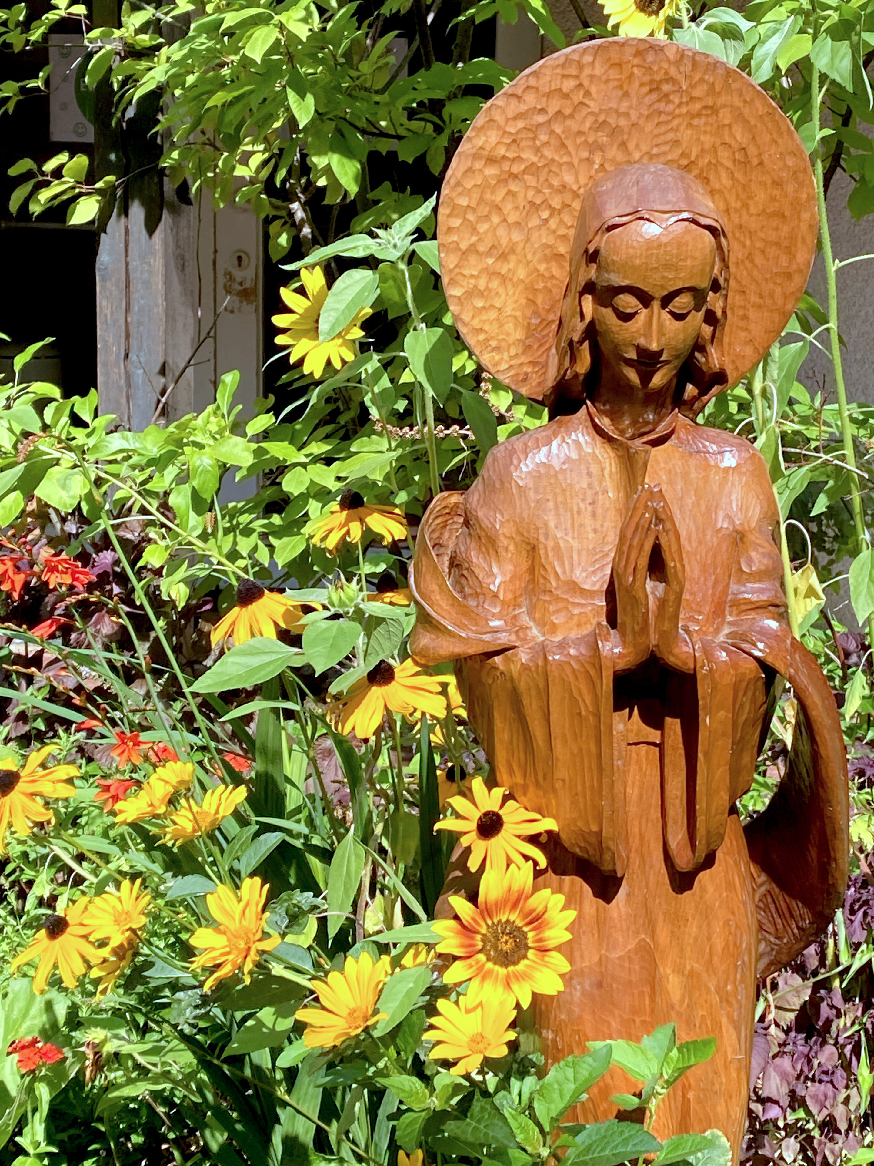 Holzfigur der betenden Gottesmutter Maria im Garten, umgeben von gelben Sonnenblumen.